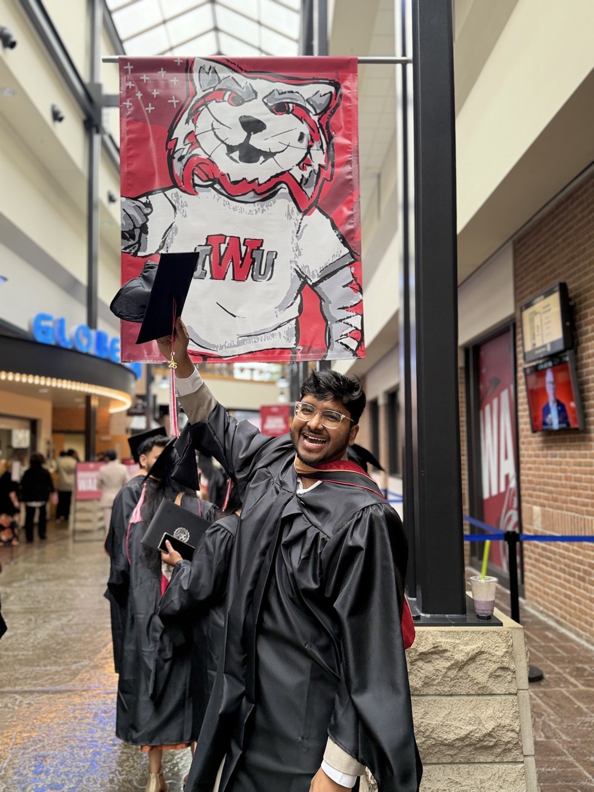 Bharath Chandra celebrating in graduation regalia beneath an Indiana Wesleyan University banner